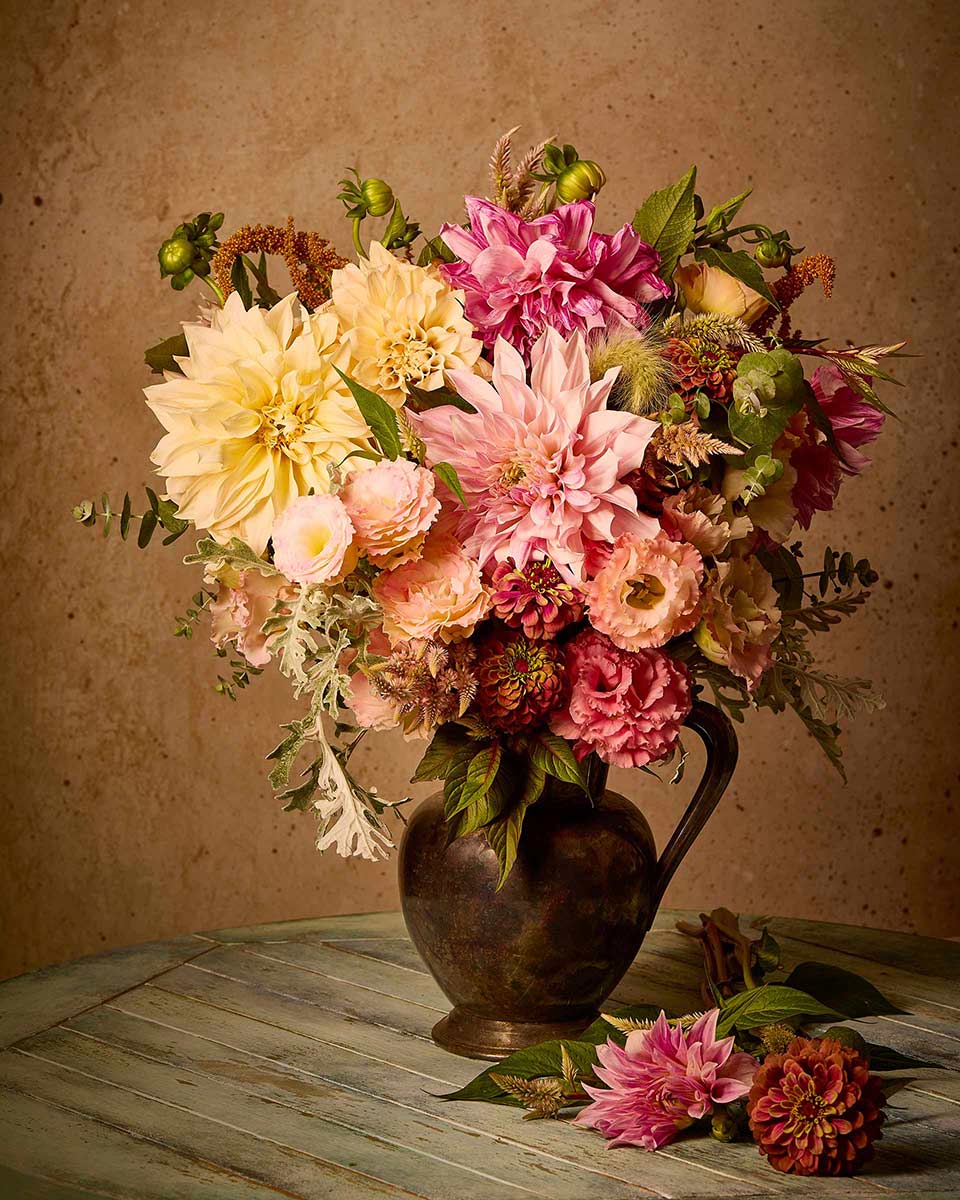 A rustic metal pitcher holds a vibrant bouquet of pink, yellow, and peach flowers on a round wooden table, mirroring the Dahlia Obsession. The backdrop features a textured beige wall like a giclee fine art print, with flower stems and leaves scattered around.