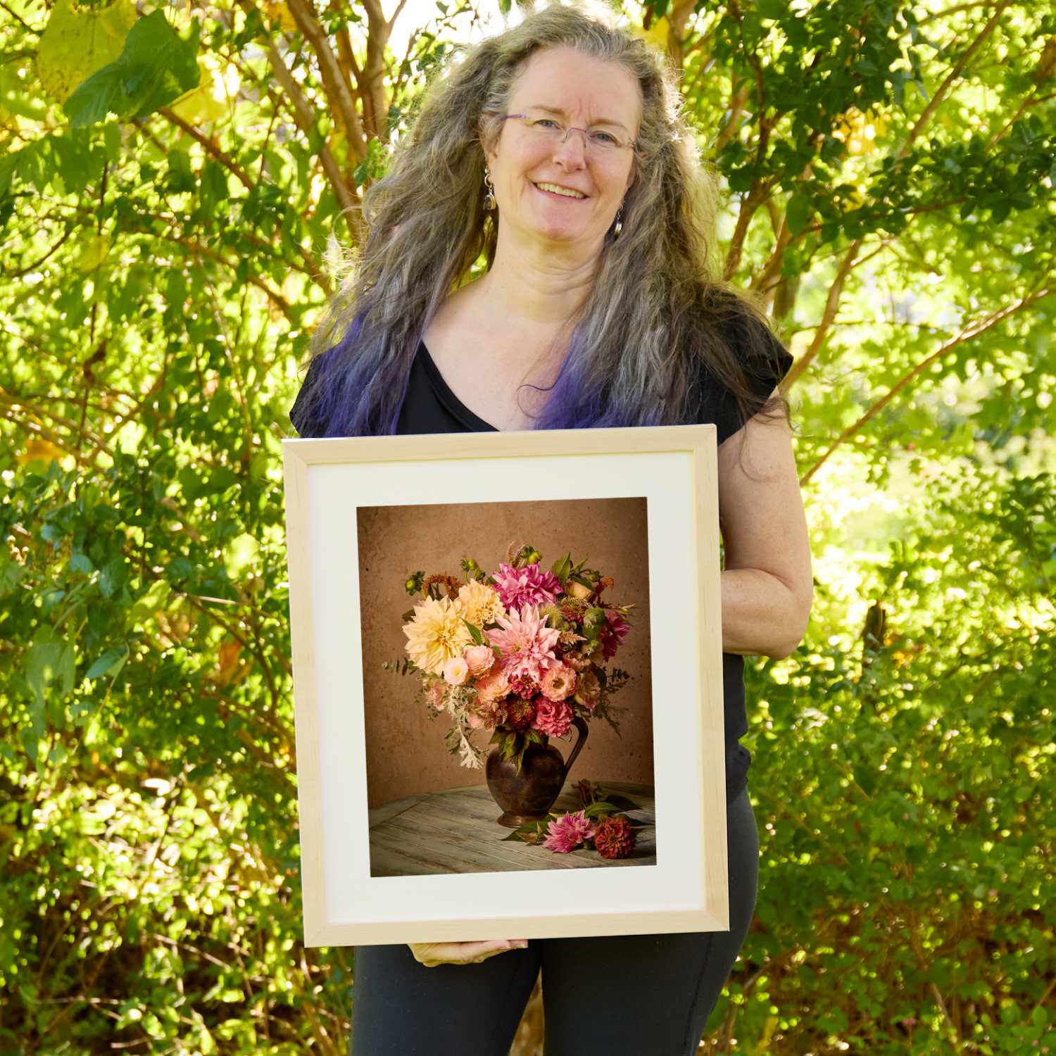 Christina Peters holding Dahlia Obsession Wall Art Print, a still life of peach, pink and burgundy dahlias in a dark bronze pitcher on a weathered wooden table, framed in natural wood with a white mat. She is standing outdoors in front of sunlit green trees and leafy shrubs.