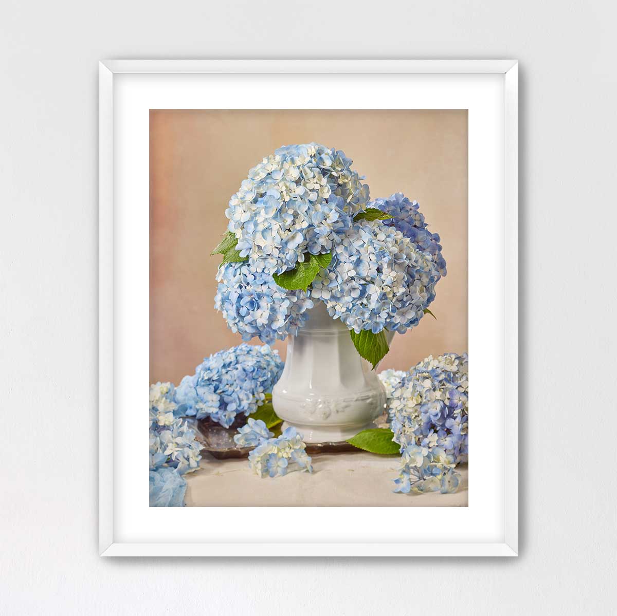 A white vase filled with blue hydrangea flowers sits on a table, surrounded by additional blue hydrangea flowers.