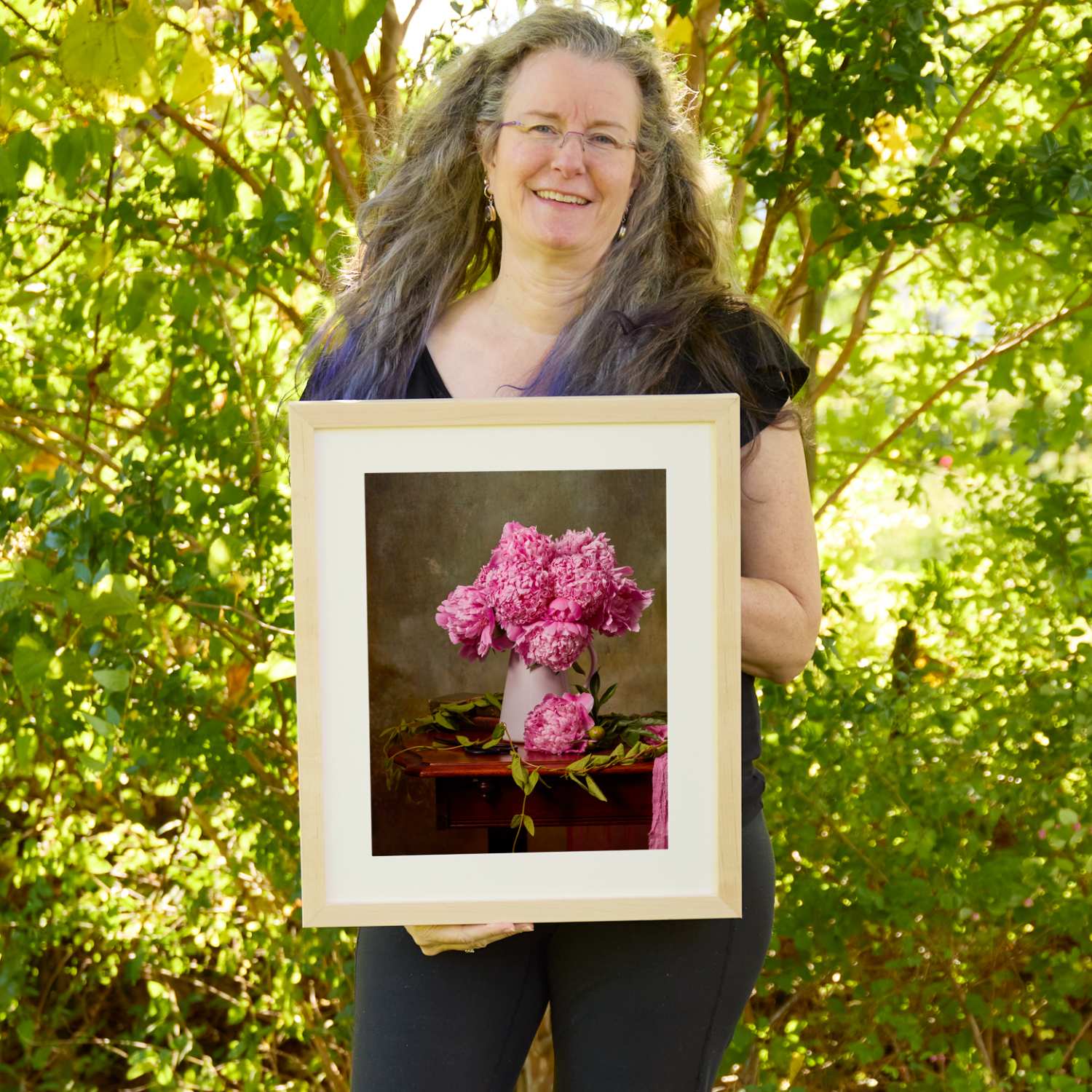 Christina Peters holding The Peony Sisters Wall Art Print, a still life of lush magenta-pink peonies in a white pitcher on a dark wooden table, with a single peony and green leaves at its base against a dark moody backdrop, framed in natural wood with a white mat. She is standing outdoors in front of sunlit green trees and leafy shrubs.