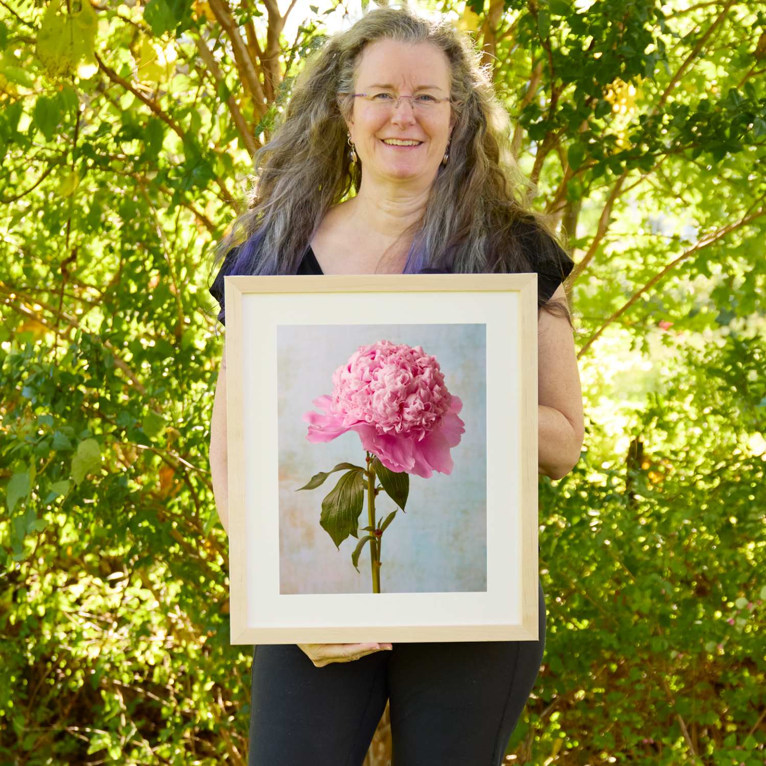 Christina Peters holding Miss Stoic Peony Wall Art Print, a single ruffled pink peony on a tall green stem with leaves against a soft pale blue textured backdrop, framed in natural wood with a white mat. She is standing outdoors in front of sunlit green trees and leafy shrubs.