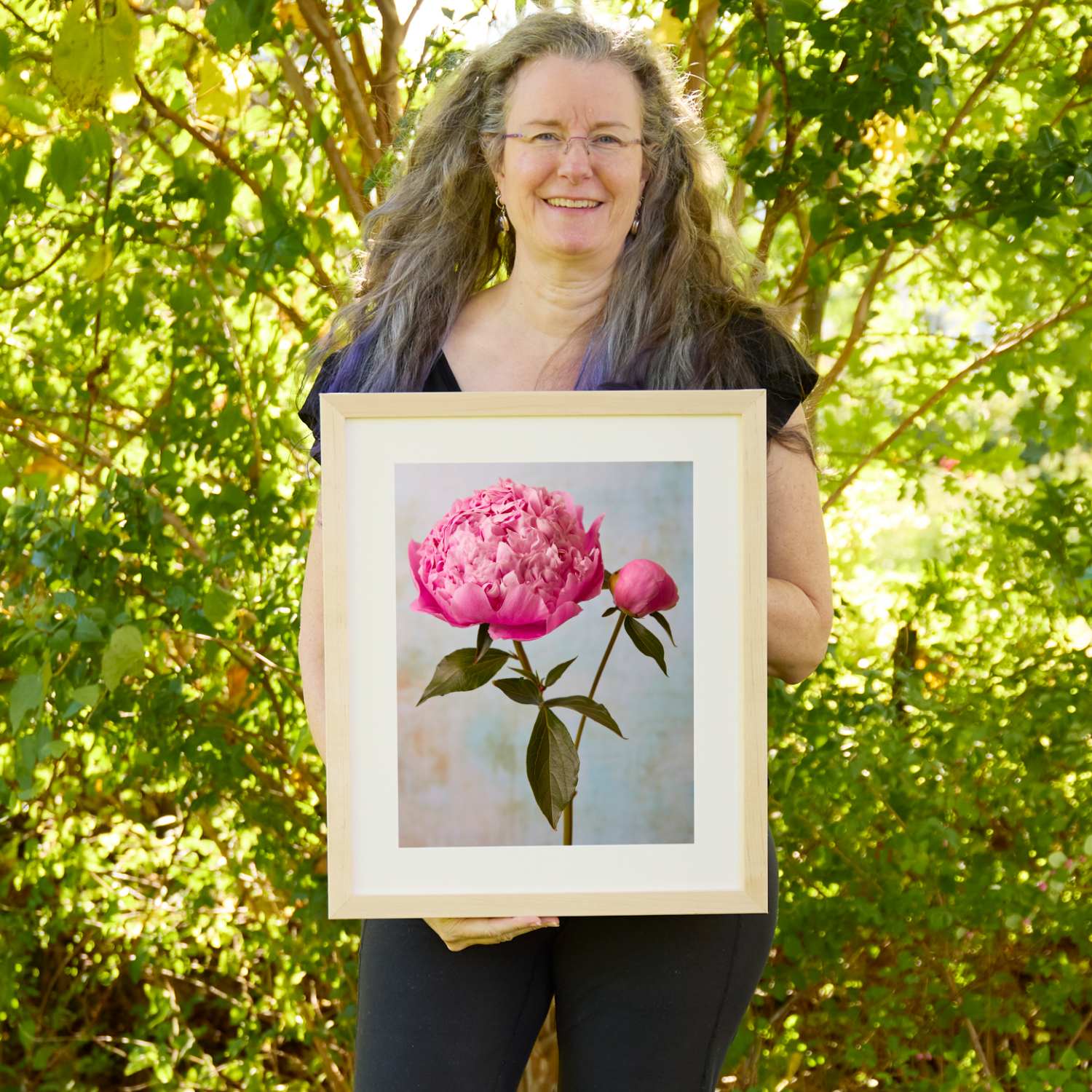 Christina Peters holding Miss Sissy Peony Wall Art Print, a full hot-pink peony and a closed bud on green stems against a soft pale blue textured backdrop, framed in natural wood with a white mat. She is standing outdoors in front of sunlit green trees and leafy shrubs.