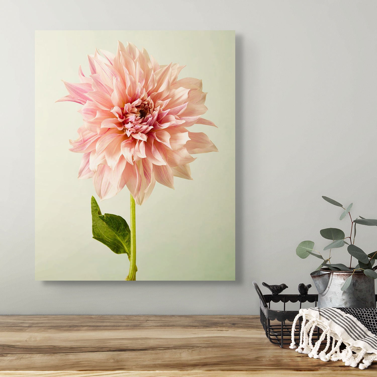 The Elegance In Pink canvas art depicting a large Café au Lait dahlia with green leaves on a light background adorns the wall. Below, a wooden table showcases a black wire basket with a white cloth and eucalyptus stems, crafting an elegant tableau.