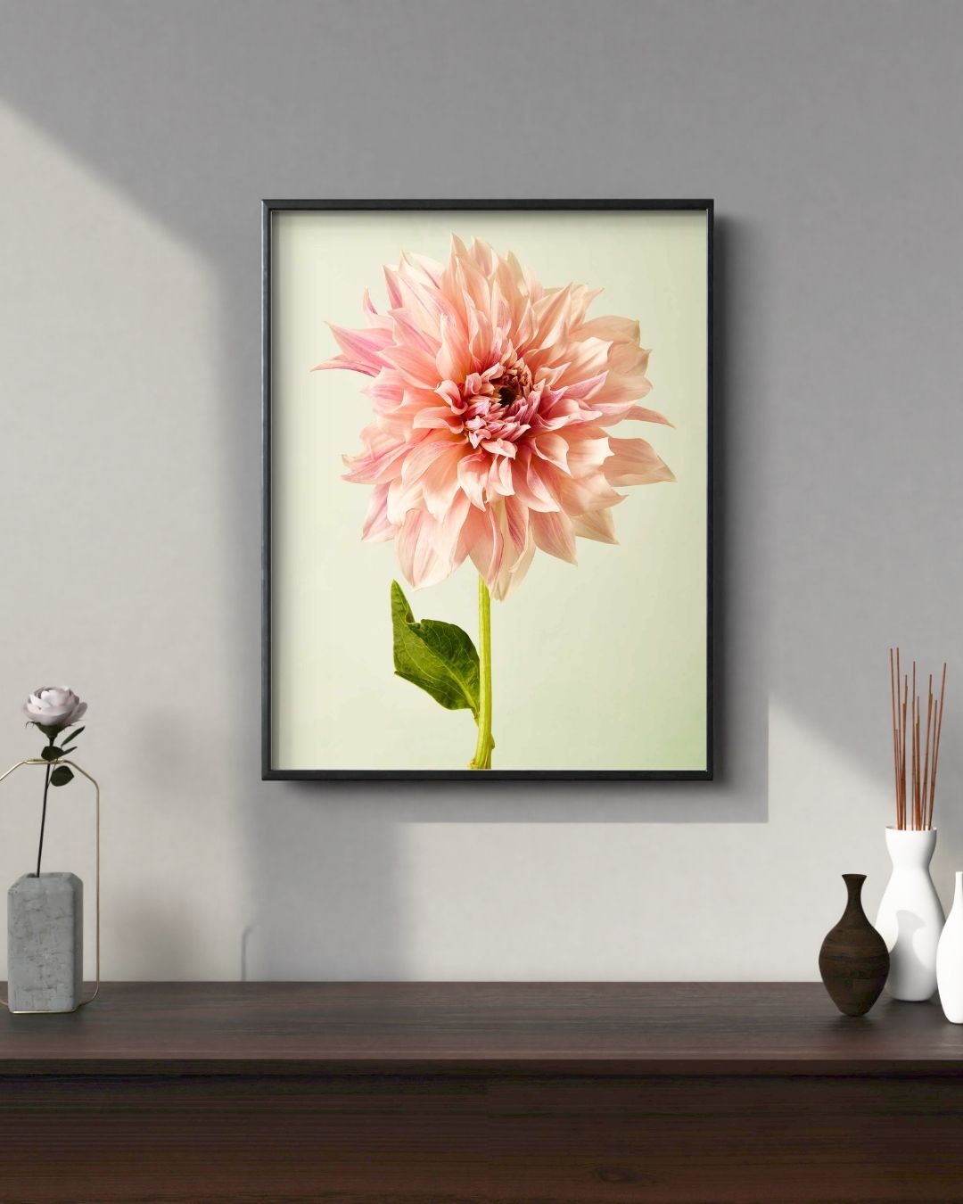 A framed picture titled Elegance In Pink featuring a large pink Café au Lait dahlia on a gray wall is complemented by a wooden shelf below, showcasing a matching dahlia in fine art giclee style, plus decorative items like two white vases and reed diffusers.