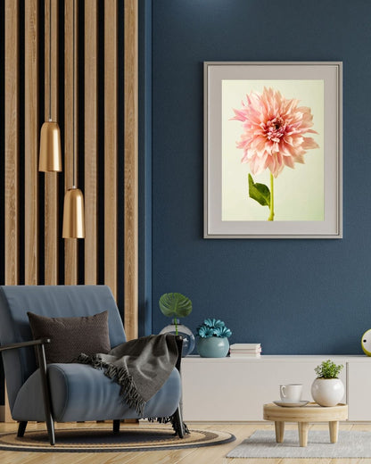 A modern living room displays Elegance In Pink, a framed painting of a large Café au Lait dahlia on a blue wall. It includes a blue armchair, gray throw, dark cushion, gold pendant lights, and wooden paneling. A small table with a plant and two white cups adds to the decor.