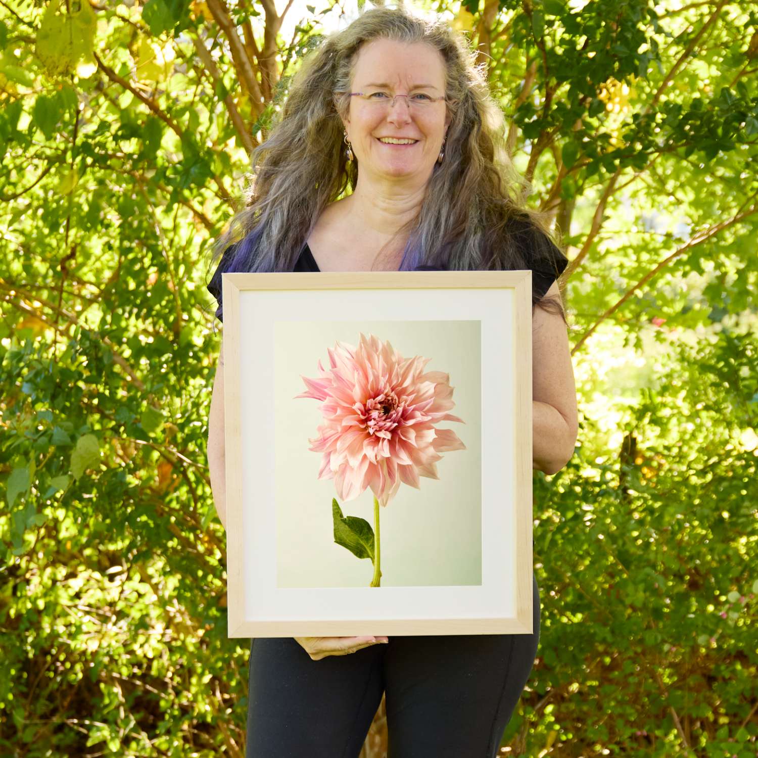 Christina Peters holding Elegance In Pink Dahlia Wall Art Print, a single blush-pink dinnerplate dahlia on a green stem against a soft pale backdrop, framed in natural wood with a white mat. She is standing outdoors in front of sunlit green trees and leafy shrubs.