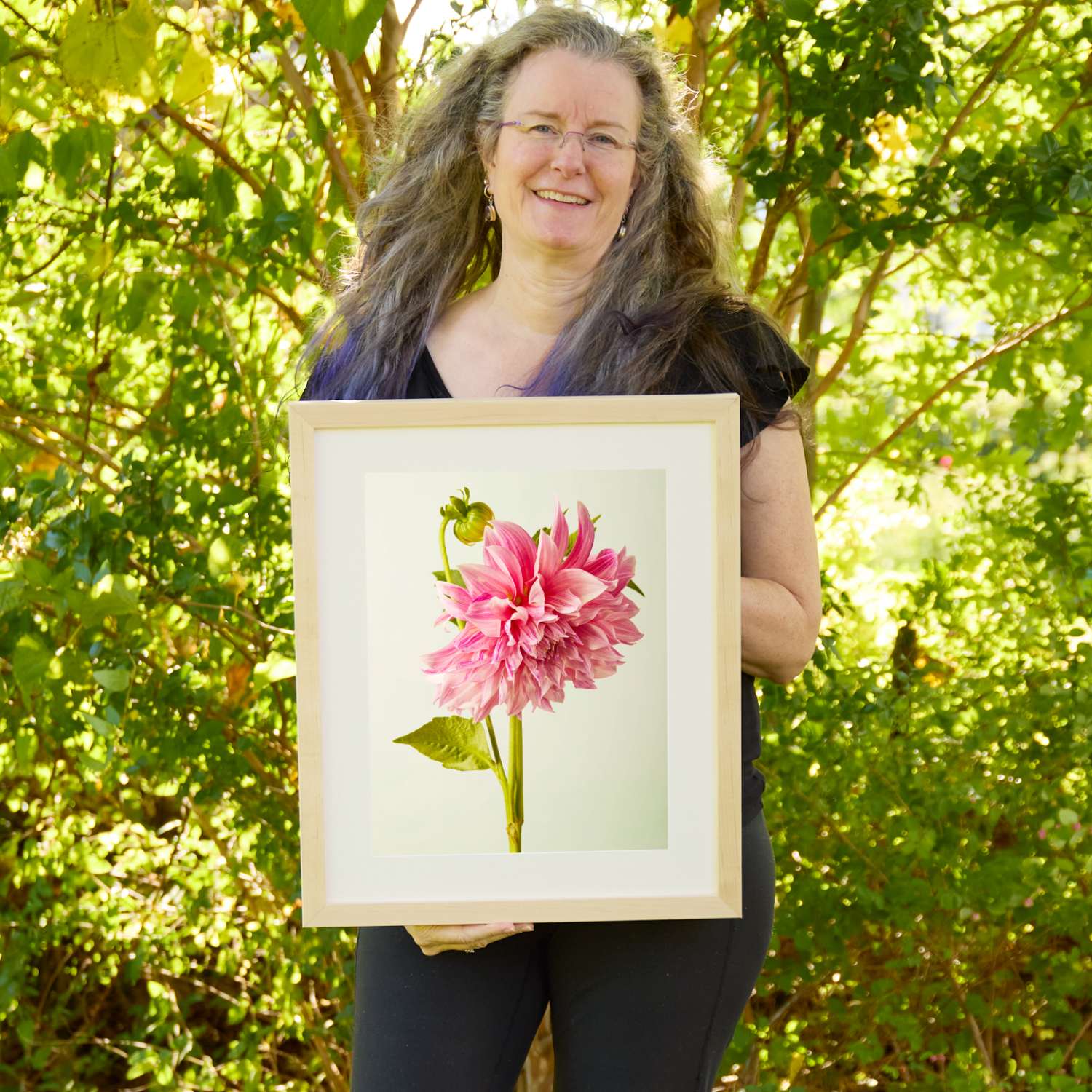 Christina Peters holding Budding Beauty Dahlia Wall Art Print, a single rose-pink dahlia in full bloom with a green bud above it on a tall stem against a soft pale backdrop, framed in natural wood with a white mat. She is standing outdoors in front of sunlit green trees and leafy shrubs.
