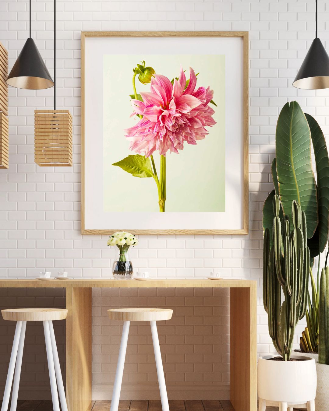 A stylish room features a wooden table with two white stools. The white brick wall showcases a Budding Beauty Dahlia paper print of a pink dahlia. Three pendant lights and potted plants, including a cactus, adorn the area. A small vase with flowers sits on the table.