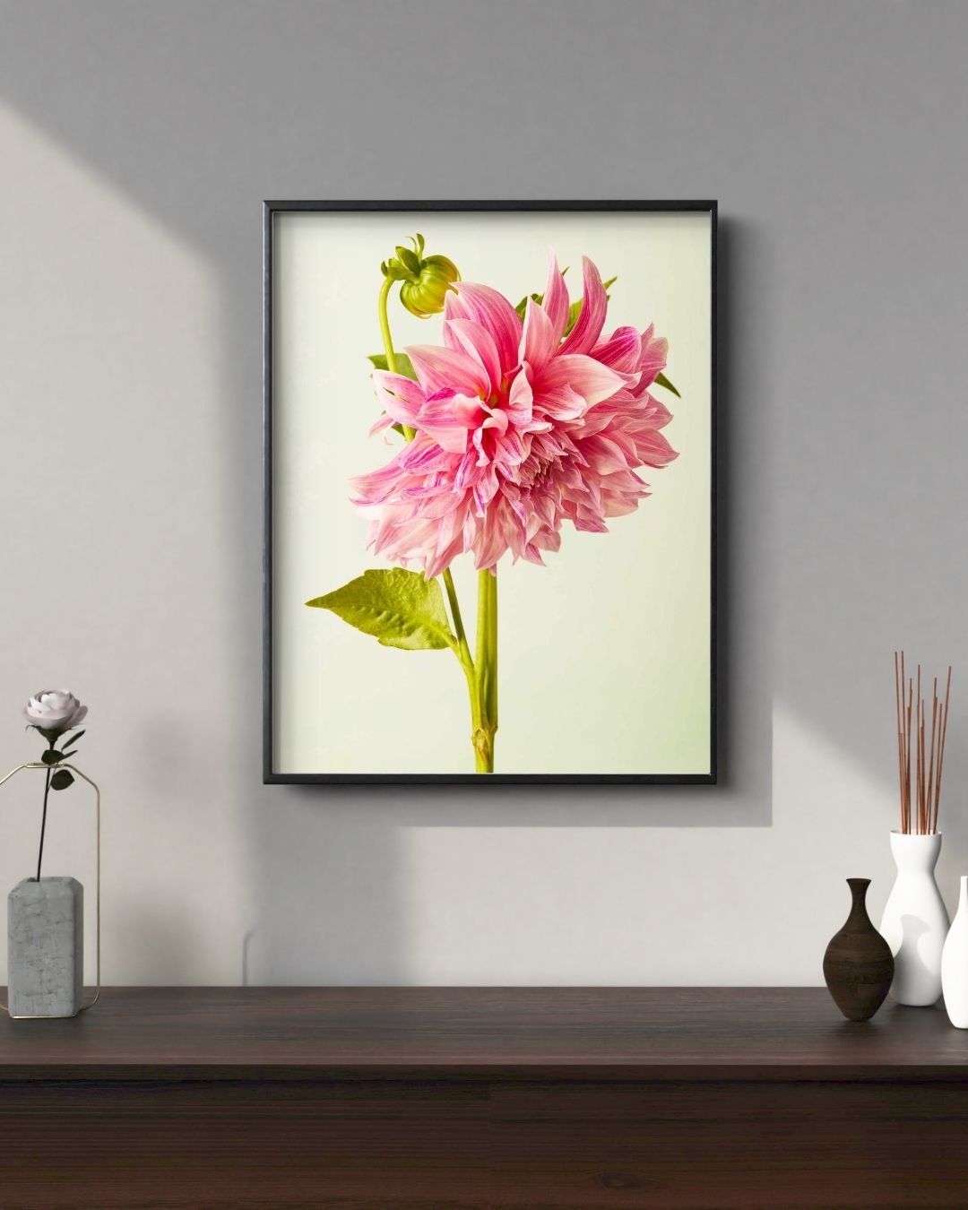 The Budding Beauty Dahlia framed art piece, printed on fine art canvas, adorns a gray wall. Below it, a dark wooden table features a small vase with a flower, two white vases, and reed diffusers. Soft light casts gentle shadows on the wall.