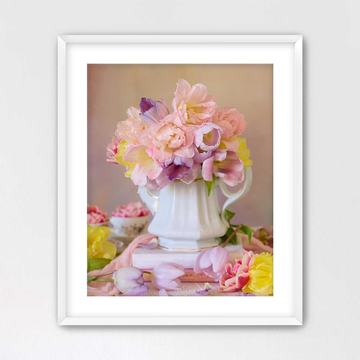 A white vase filled with a vibrant bouquet of pink and yellow flowers sits on a pink surface, surrounded by additional flowers.