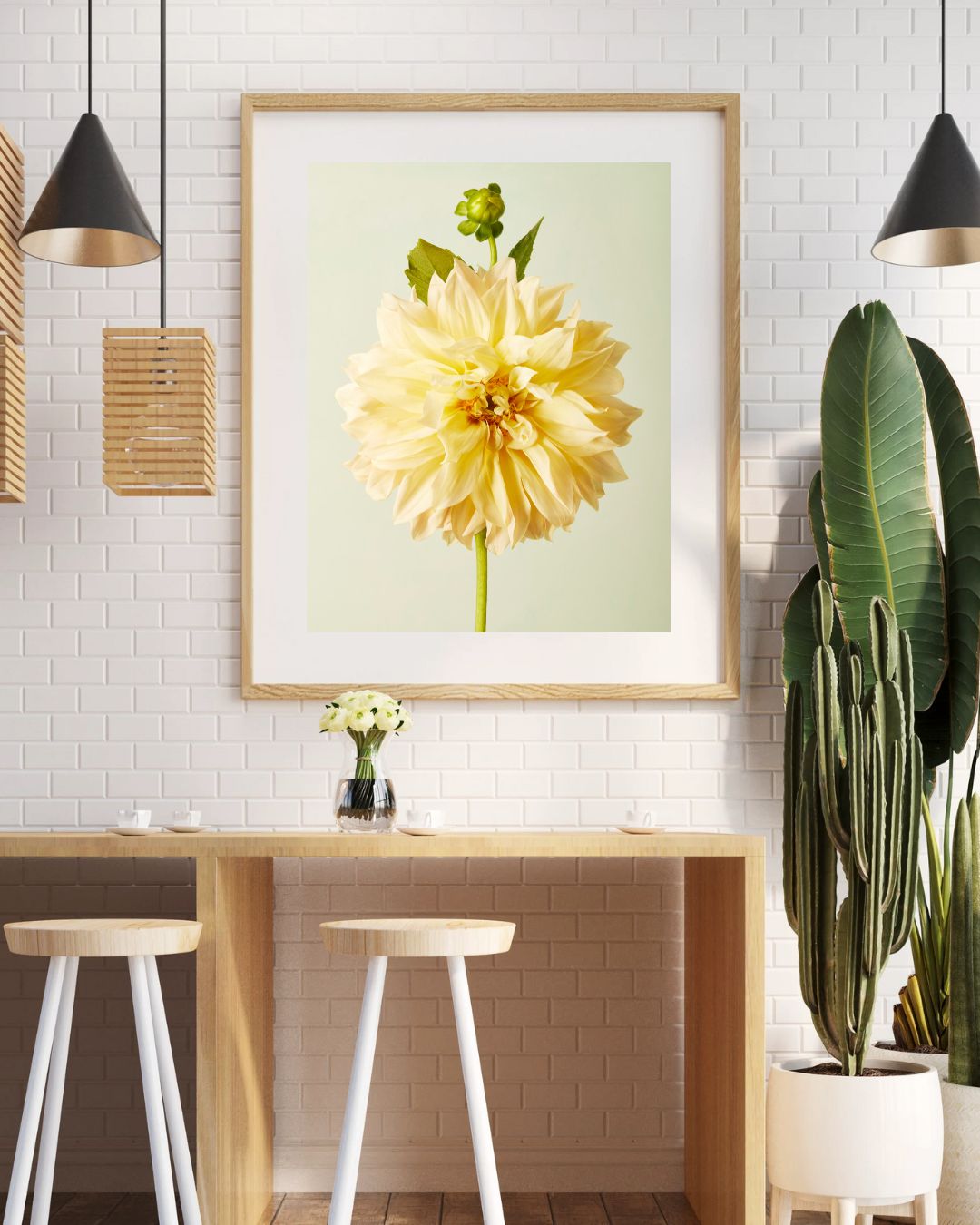 A modern kitchen with a wooden table and two white stools showcases the Champagne Petals fine art giclee of a Café au Lait dahlia on the white brick wall. Pendant lights, a potted cactus, and a small vase of flowers enhance the ambiance.