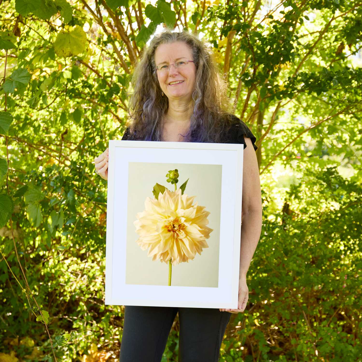 Christina Peters holding Champagne Petals Dahlia Wall Art Print, a single creamy champagne-yellow dinnerplate dahlia with a bud above it on a green stem against a soft pale backdrop, framed in white with a white mat. She is standing outdoors in front of sunlit green trees and leafy shrubs.