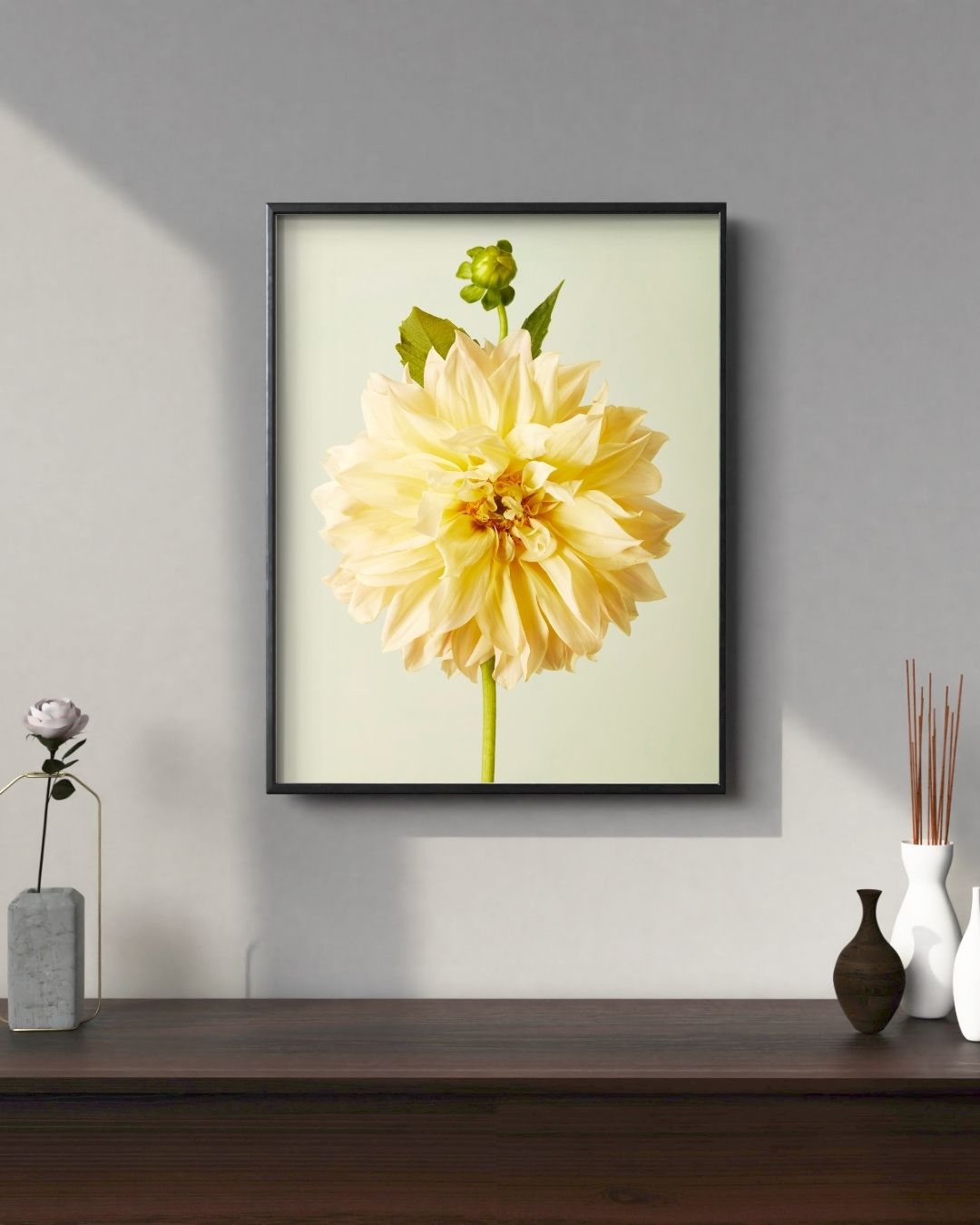 The Champagne Petals canvas wall art showcases a large pale yellow Café au Lait dahlia with a bud and green leaves, beautifully framed on a light gray wall. Below, a dark wood surface holds a small flower in a vase, a white vase, and room diffuser with reeds.