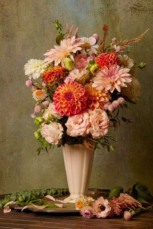 The Victorian Bouquet, in a tall cream vase, displays pink, orange, and white dahlias and roses with greenery. Scattered blossoms and greenery embellish the wooden table below against a green textured backdrop reminiscent of canvas wall art.