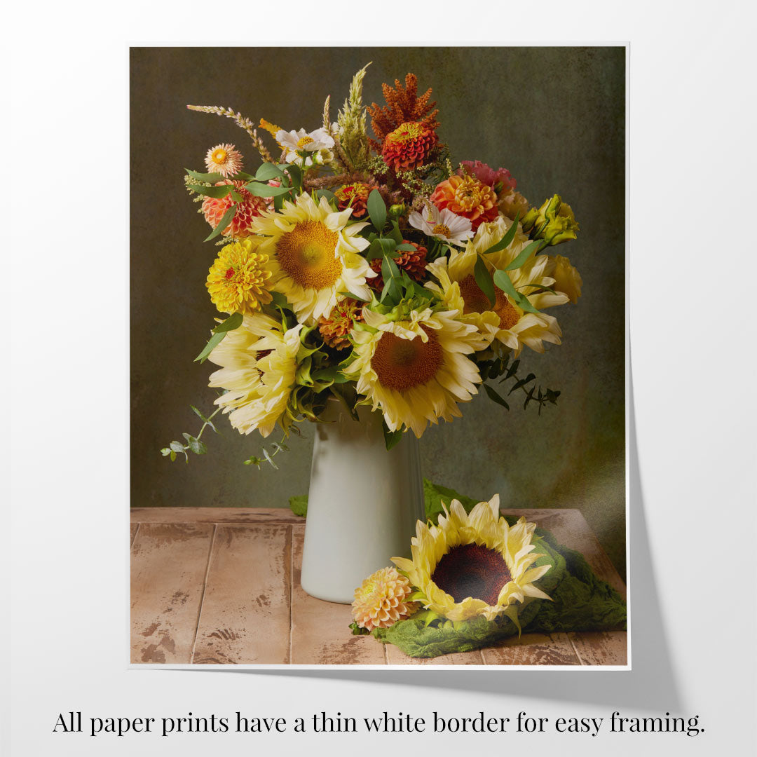 A photographic print of The Sunny Bouquet is displayed on a wooden surface, featuring vibrant sunflowers and colorful blooms. Next to it rests a single sunflower, inviting admiration. The paper edge is curled up and the print has a thin white border.