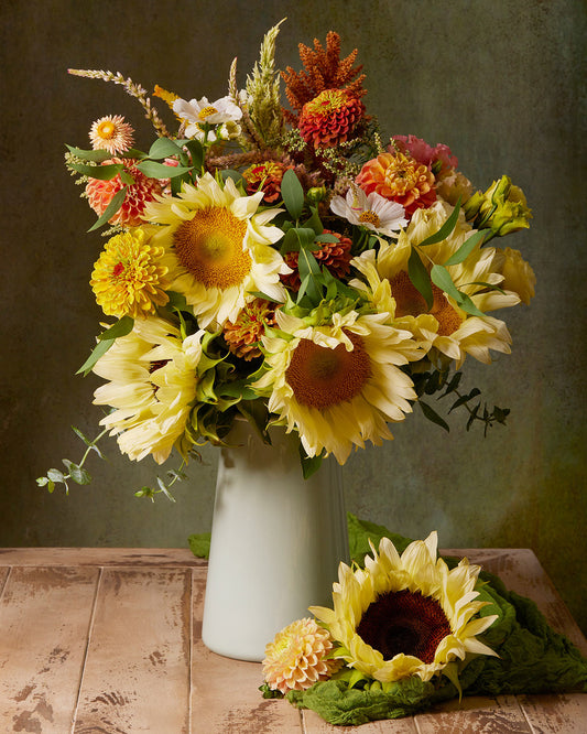 Capture the beauty of the Sunny Bouquet, featuring a vibrant arrangement of sunflowers, zinnias, and wildflowers in yellow, orange, and pink hues. Presented in a light green vase with one sunflower and a pink dahlia beside it on wood. Available as a fine art paper print or as canvas wall art.