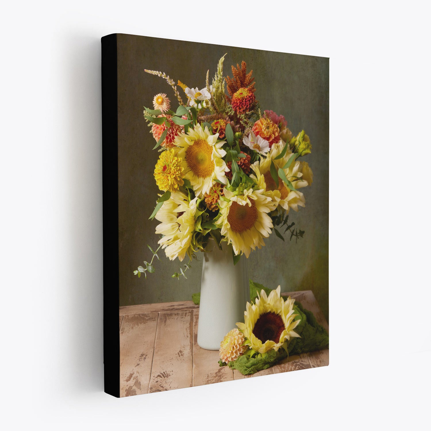 The Sunny Bouquet canvas wall art beautifully depicts a still life scene with vibrant sunflowers and wildflowers in a white vase on a wooden table, complemented by one sunflower lying beside it. A muted green background adds to the artworks serene charm. This canvas wrap has black edges.