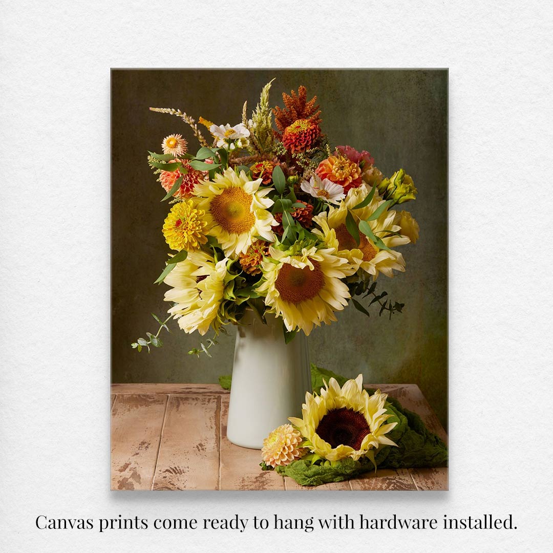 The Sunny Bouquet canvas wall art features a vibrant arrangement of sunflowers, daisies, and colorful flowers in a white vase on rustic wood, with a sunflower beside it. Comes ready to hang with hardware installed.