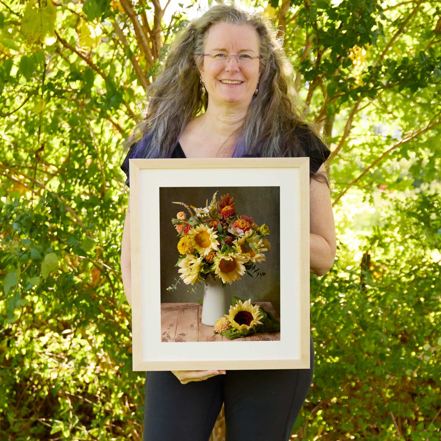 Christina Peters holding Sunny Bouquet Sunflower Wall Art Print, a still life of pale yellow sunflowers, orange dahlias and summer blooms in a white pitcher on a rustic wooden table, with a single sunflower beside it against a dark backdrop, framed in natural wood with a white mat. She is standing outdoors in front of sunlit green trees and leafy shrubs.