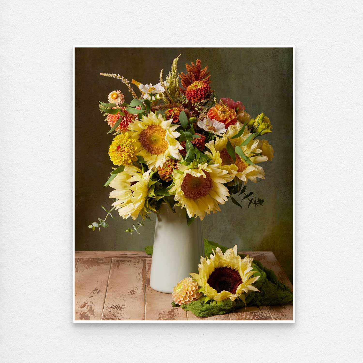 The Sunny Bouquet, showcasing vibrant sunflowers and autumn flowers in orange and yellow, sits elegantly in a pale green vase atop a wooden table against a dark green backdrop, reminiscent of fine art, with a fallen sunflower enhancing the charm. Print shows with a thin white border for easy framing.