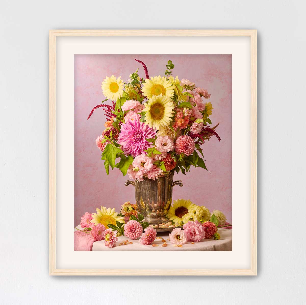 A framed photograph of a vase filled with a vibrant bouquet of yellow and pink flowers, placed on a table against a pink background.