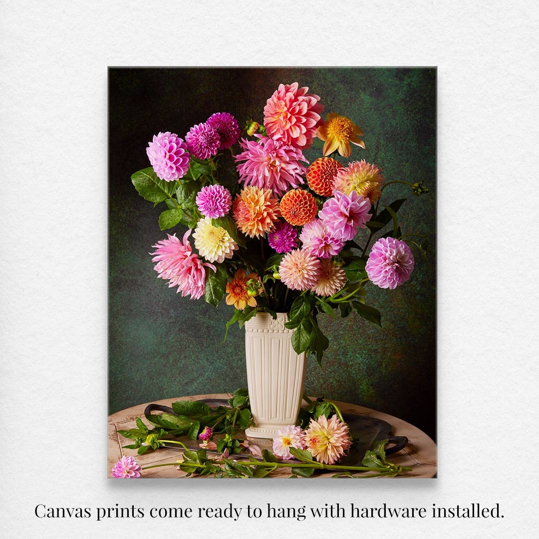 The Garden's Grand Finale canvas features a vibrant still life of pink, orange, purple, and yellow dahlias in a white vase against a dark green textured background. Scattered leaves accentuate the base, making it ideal for canvas wall art. A caption beneath states the canvas print comes ready to hang.