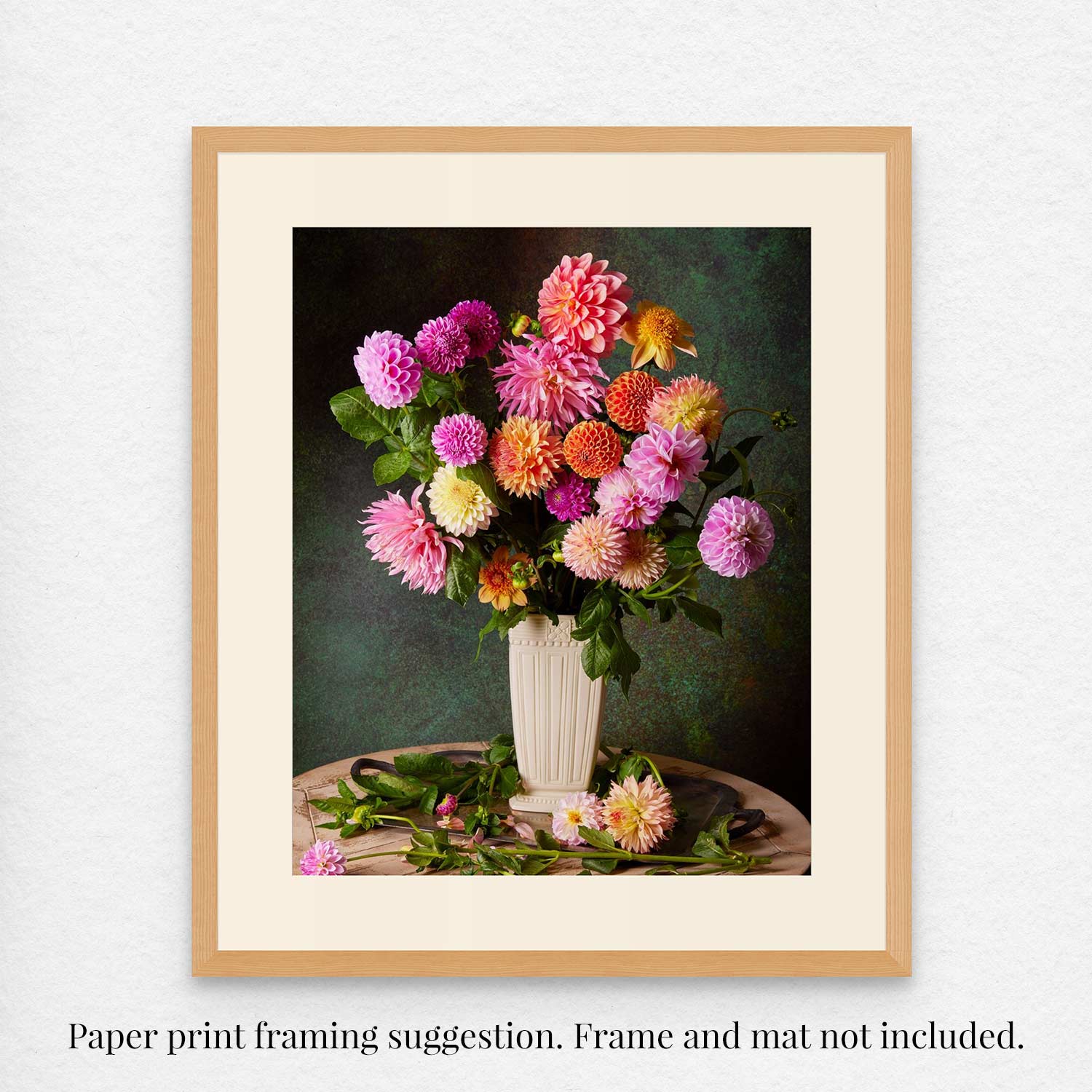 Garden's Grand Finale is a framed fine art paper print showcasing vibrant pink, orange, and yellow dahlias in a white vase on a dark green background, with extra blossoms and leaves scattered below. Frame and mat are not included.