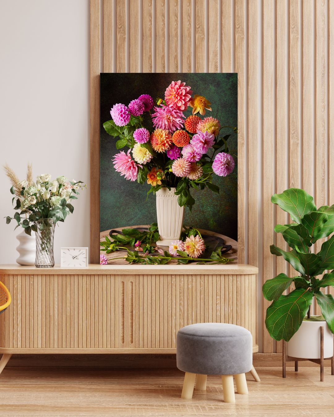 A modern interior features Garden's Grand Finale canvas art, showcasing vibrant pink and orange dahlias in a vase on a wooden wall. A small plant, white flowers in another vase, a clock, and a gray cushioned stool complete the decor.