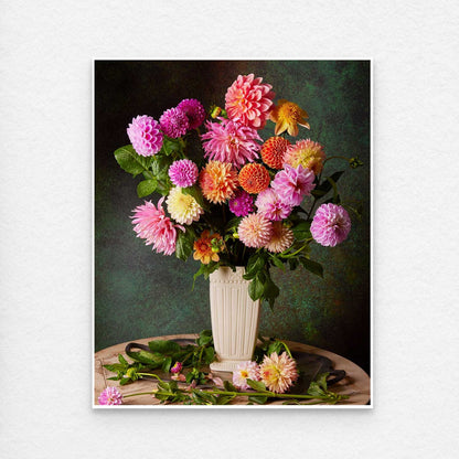 The Garden's Grand Finale features a vase brimming with vibrant multicolored dahlias—pink, orange, yellow, purple—on a round wooden table against a dark green backdrop. Scattered leaves and flowers enhance this scene. This print is showing a think white border to make for easy framing.