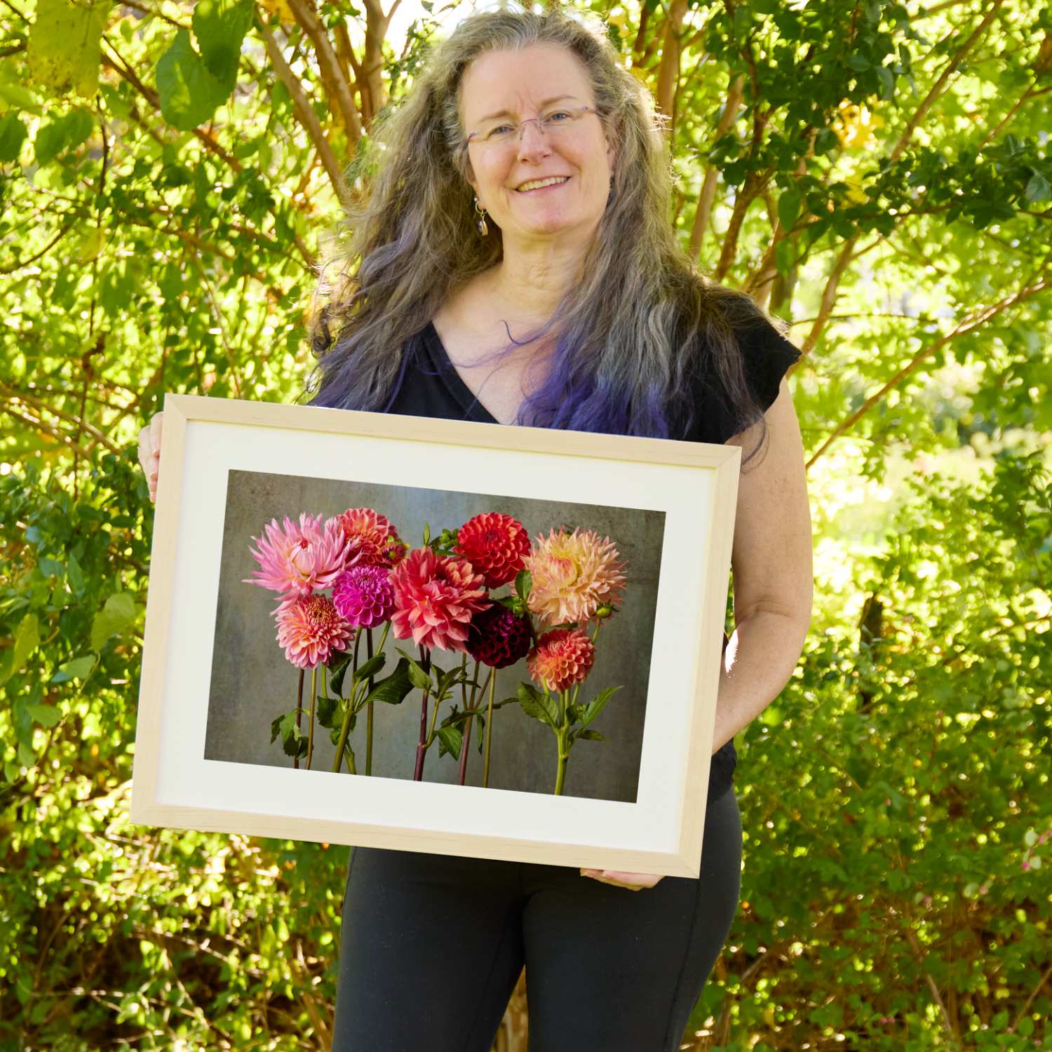 Christina Peters holding Summer's Chorus Dahlia Fine Art Print, a row of pink, coral, red, burgundy and peach dahlias standing on long stems against a weathered gray backdrop, framed in natural wood with a white mat. She is standing outdoors in front of sunlit green trees and leafy shrubs.