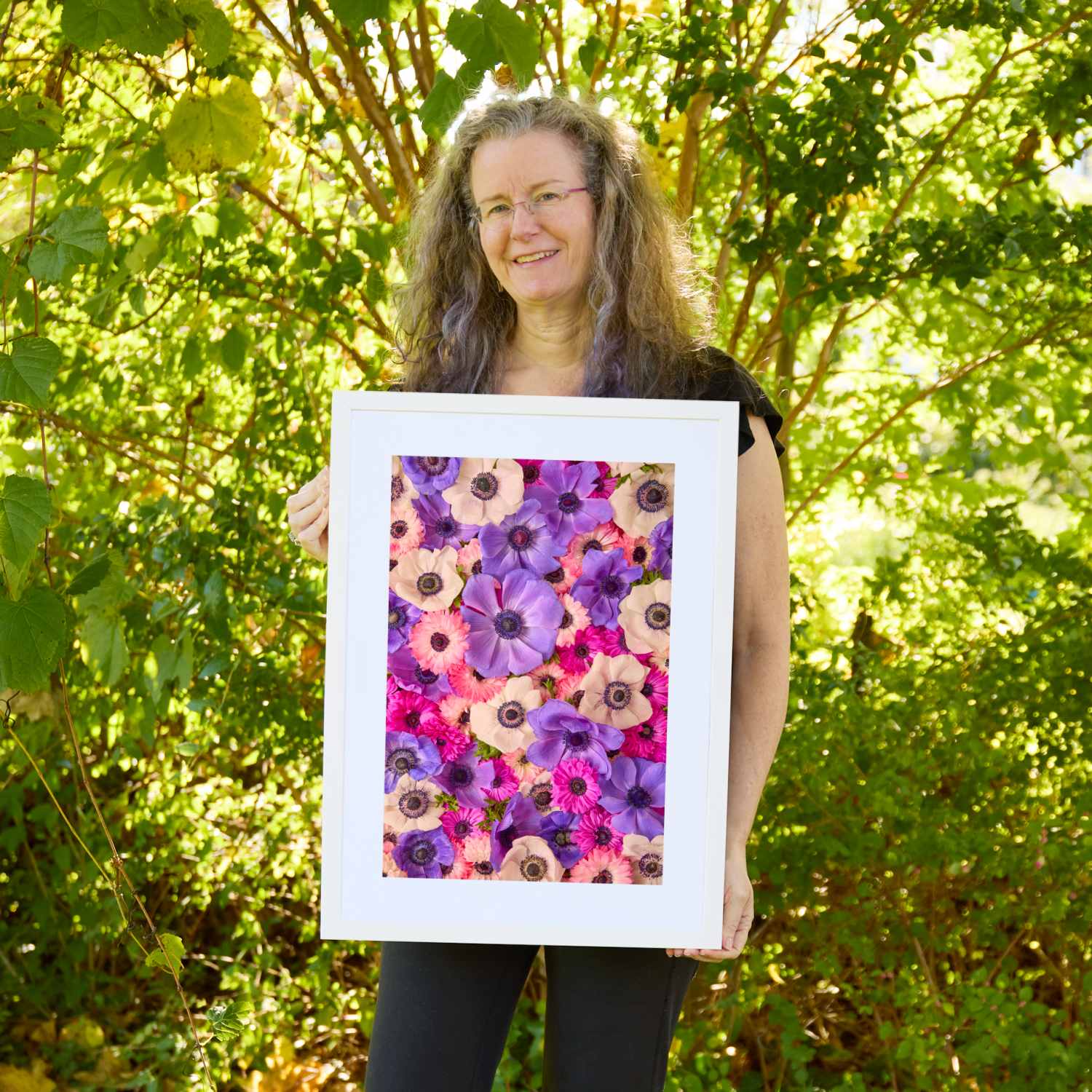 Christina Peters holding Purple Passion Anemone Wall Art Print Vertical, a vertical close-up of purple, magenta, pink and white anemones packed together, framed in white with a white mat. She is standing outdoors in front of sunlit green trees and leafy shrubs.