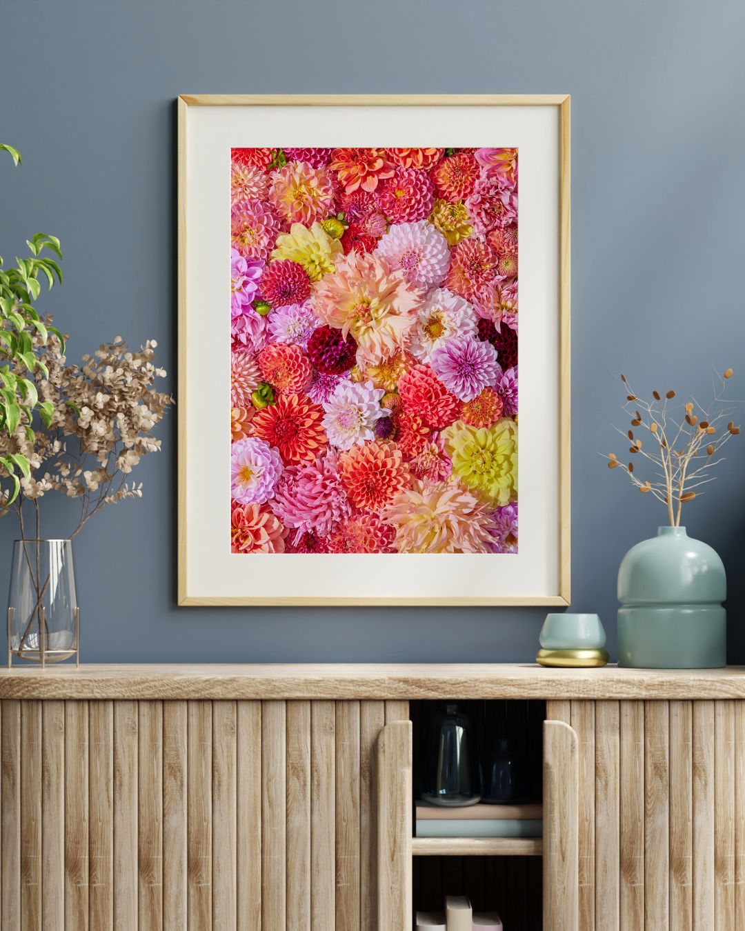 The Blooming Radiance floral canvas, showcasing vibrant flowers, hangs on a blue wall. Below, a wooden cabinet with decorative vases and dried plants enhances the lively dahlia art piece, adding charm to the room.