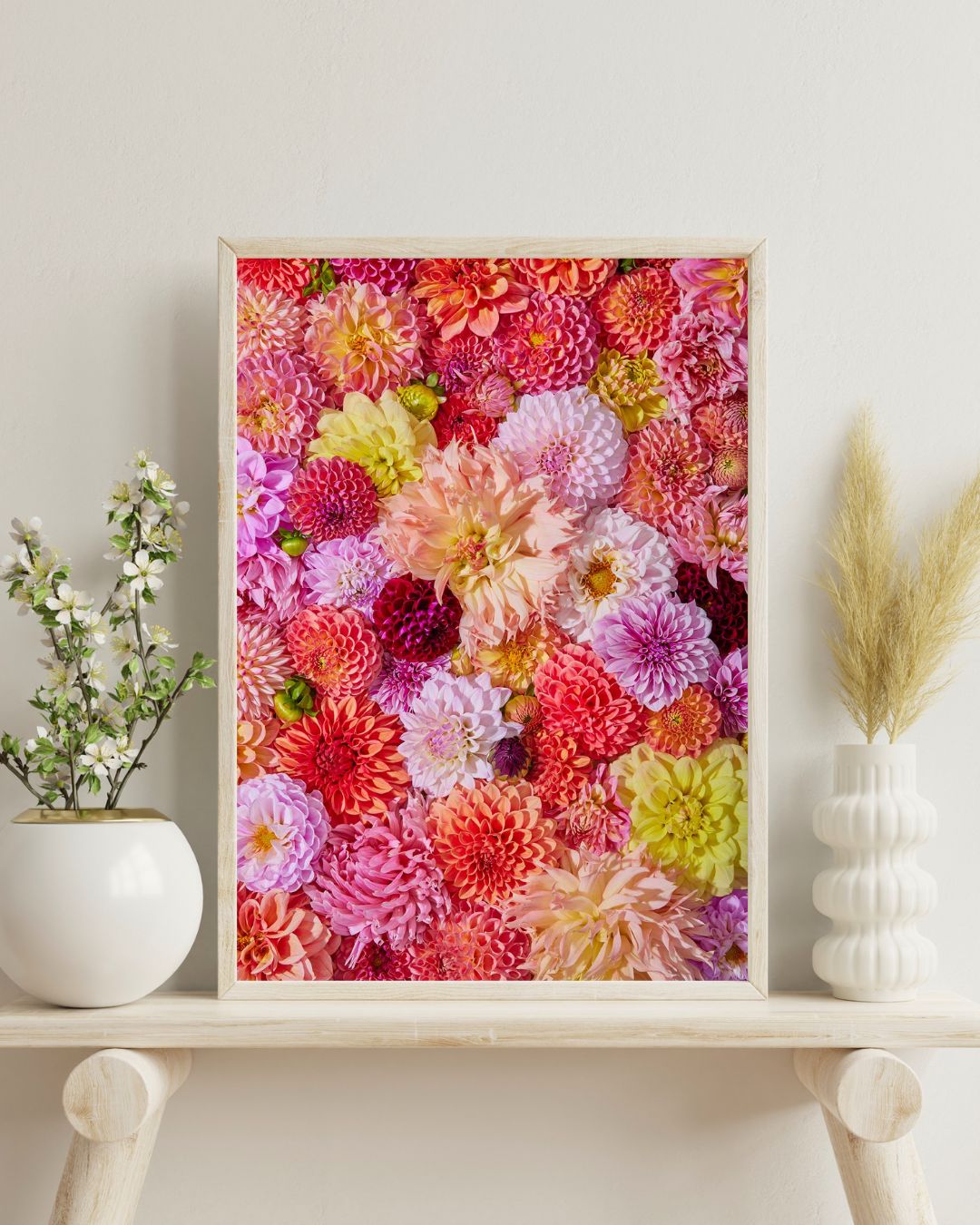 Blooming Radiance, a beautifully framed dahlia photograph, hangs on a white wall. Below, a white shelf holds vases with small white flowers and pampas grass, adding a natural touch to the decor.