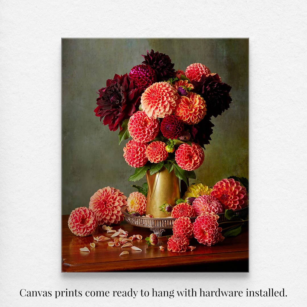 The Late Summer Glow canvas wall art features vibrant red, orange, pink, and burgundy dahlias in a brass coffee pot on a muted backdrop, with scattered petals on the wooden surface below. Ready to hang with installed hardware.