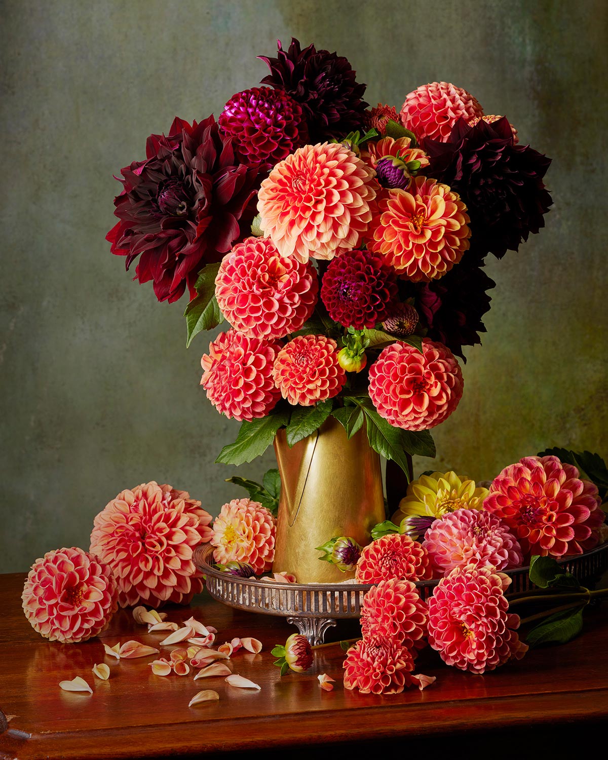 The Late Summer Glow features a vibrant dahlia arrangement in red, orange, pink, and maroon within an antique brass coffee pot. Scattered petals on the wooden table and a textured background add an elegant touch.