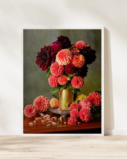 A framed canvas print of Late Summer Glow captures vibrant red and pink dahlia blooms in a brass coffee pot, with scattered petals on wood against a muted green backdrop. The canvas is resting on a light wood floor beside a white wall.