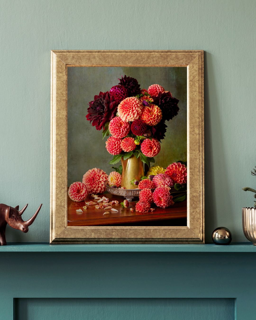 The Late Summer Glow canvas wall art framed, featuring vibrant pink and dark red dahlias in a brass coffee pot, is elegantly displayed on a teal mantel. It’s flanked by a small rhino figurine and other decorative items against a soft green wall, creating a stunning arrangement.