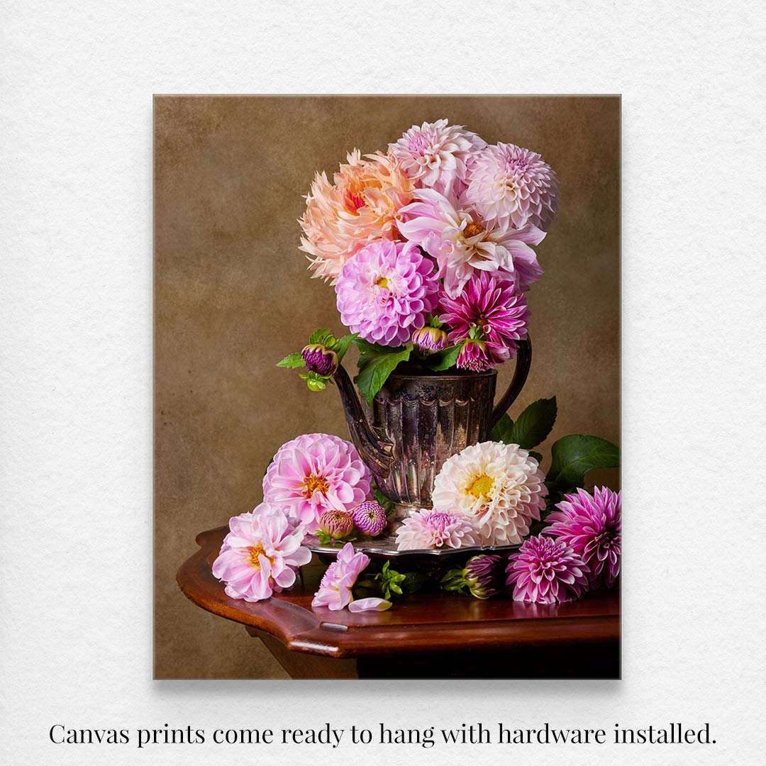 Dahlia Tea Party features pink and peach dahlias in a silver pitcher antique teapot, with scattered blooms on a wooden table. The warm brown background enriches this canvas wall art, which comes ready to hang with hardware installed.