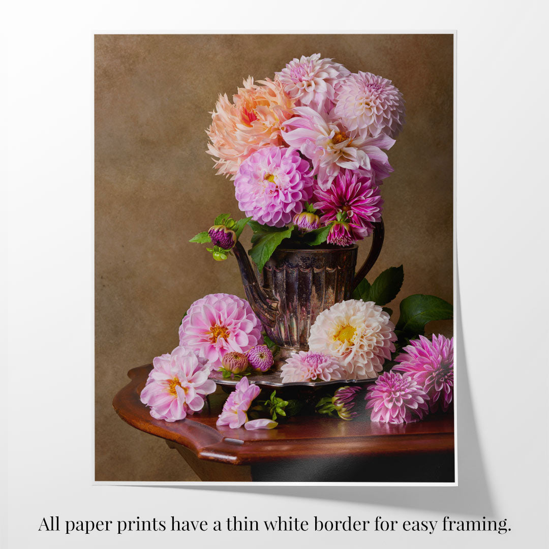 The Dahlia Tea Party paper print showcases a still life with a rustic teapot holding pink and peach dahlia blooms on a wooden table, surrounded by scattered flowers and petals. Its thin white border makes it ideal for easy framing and display.