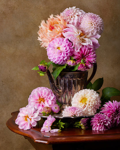 The Dahlia Tea Party features a rustic metal antique silver teapot brimming with pink and peach dahlias on a wooden table, with scattered blooms all around the base.