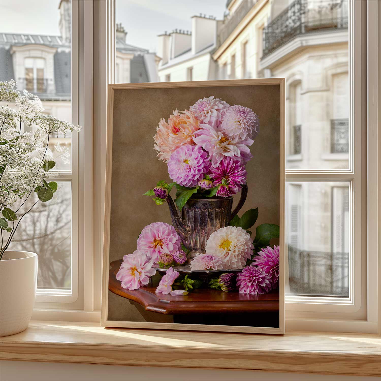 The Dahlia Tea Party canvas shown framed on a wooden windowsill. Sunlight illuminates the canvas, while a white pot with delicate plants sits nearby, overlooking the buildings outside.