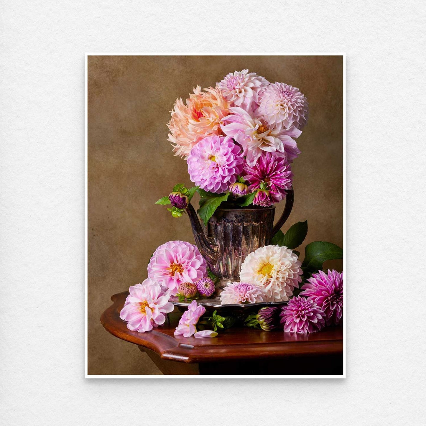 Print of Dahlia Tea Party features a vintage metal teapot filled with pink and peach dahlias, placed on a wooden table against a muted brown backdrop. Additional blooms and green leaves are artfully arranged at the base. All paper prints come with a thin white border.