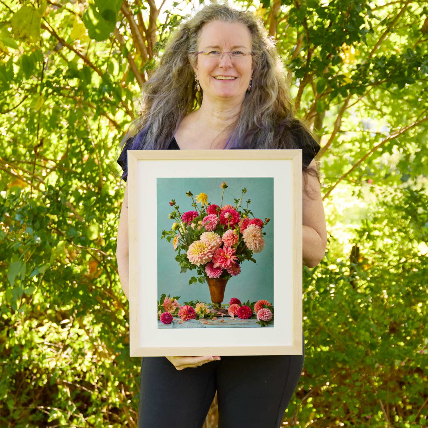 Christina Peters holding Summer's Farewell Dahlia Wall Art Print, a still life of pink, coral and peach dahlias in a terracotta vase, with loose blooms on the table against a soft teal backdrop, framed in natural wood with a white mat. She is standing outdoors in front of sunlit green trees and leafy shrubs.