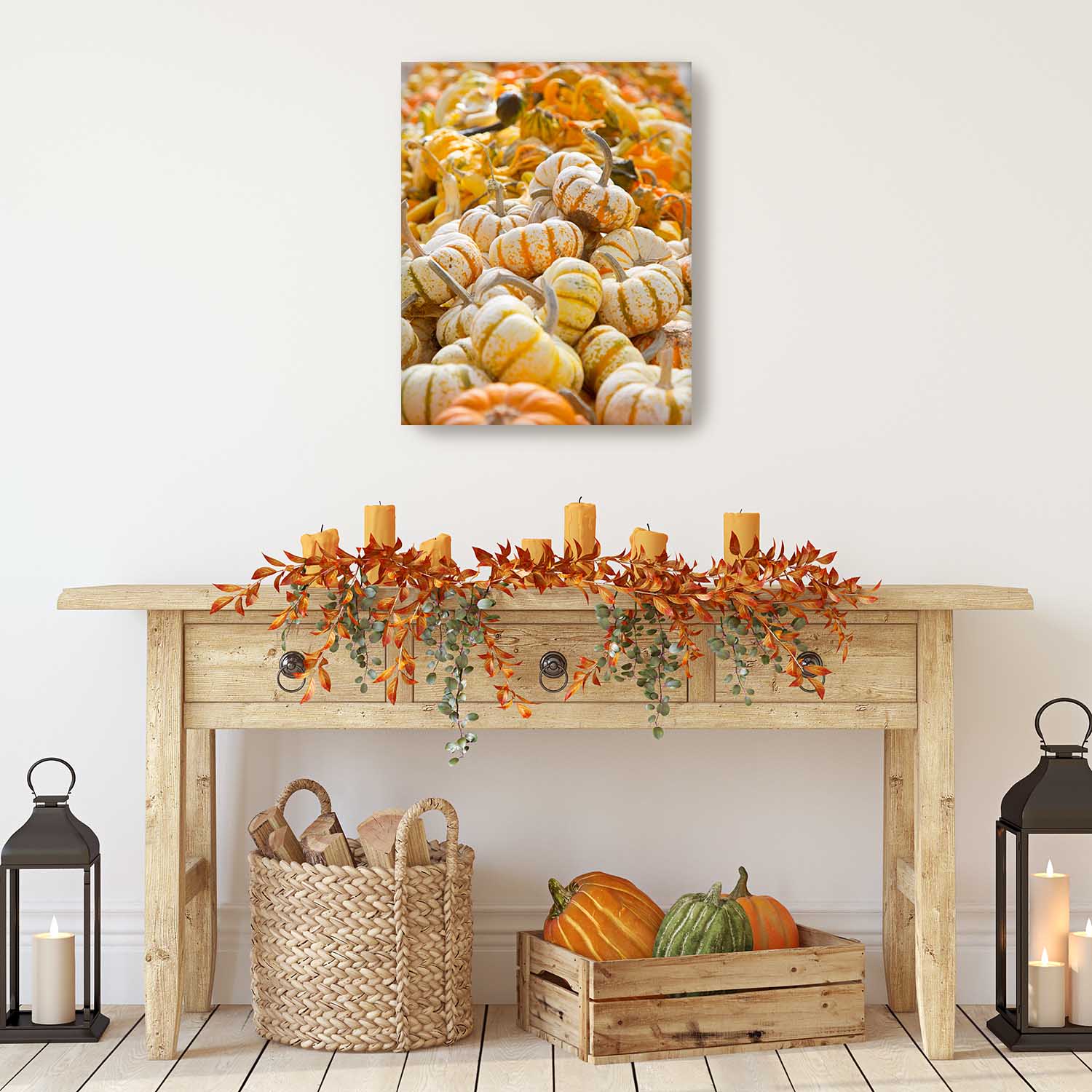 Decorative setup with pumpkins, candles, and a basket on a wooden table against a white wall with the canvas of Harvest Abundance on the wall