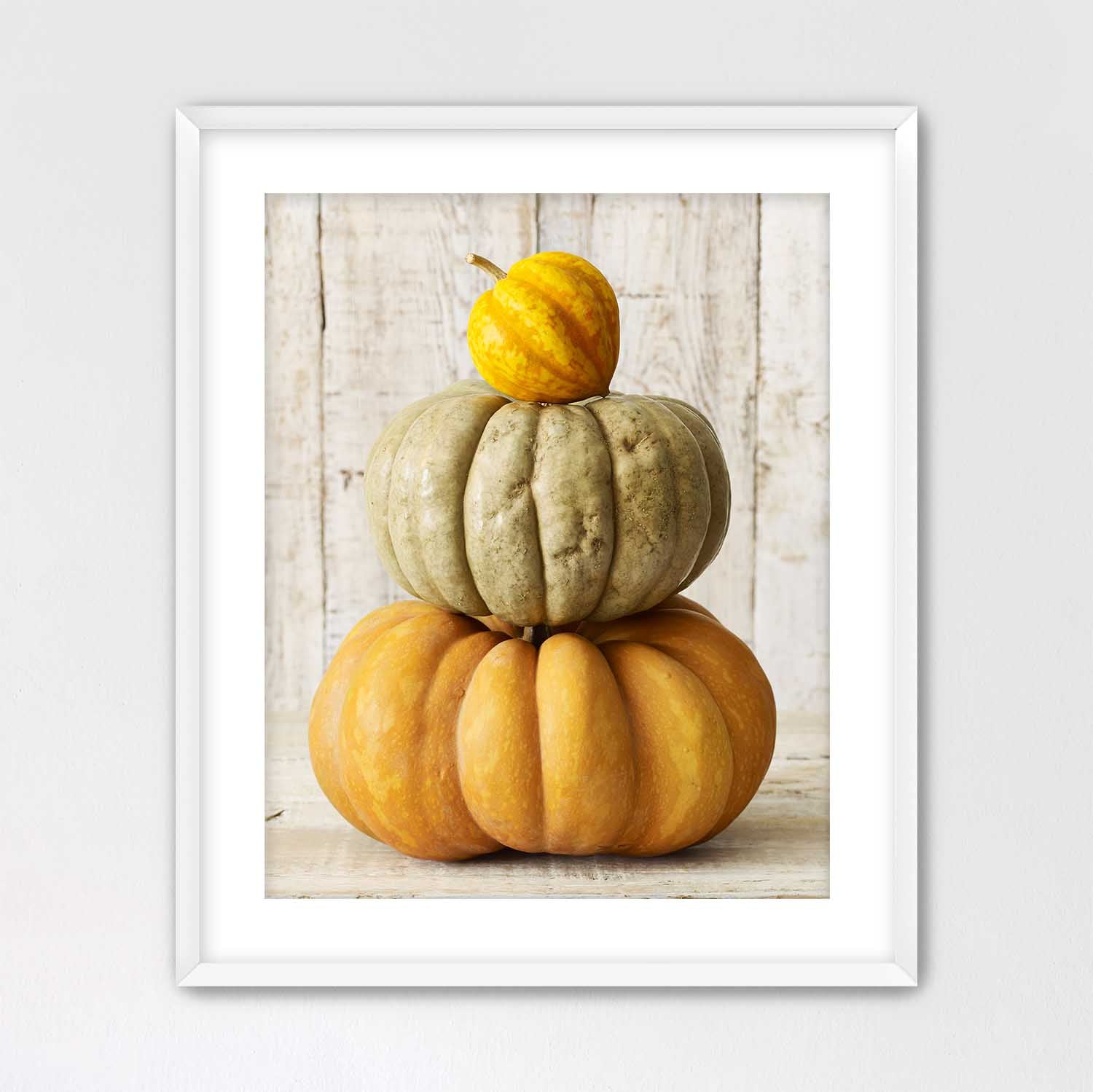 White framed print with a white mat board of three stacked pumpkins on a wooden surface