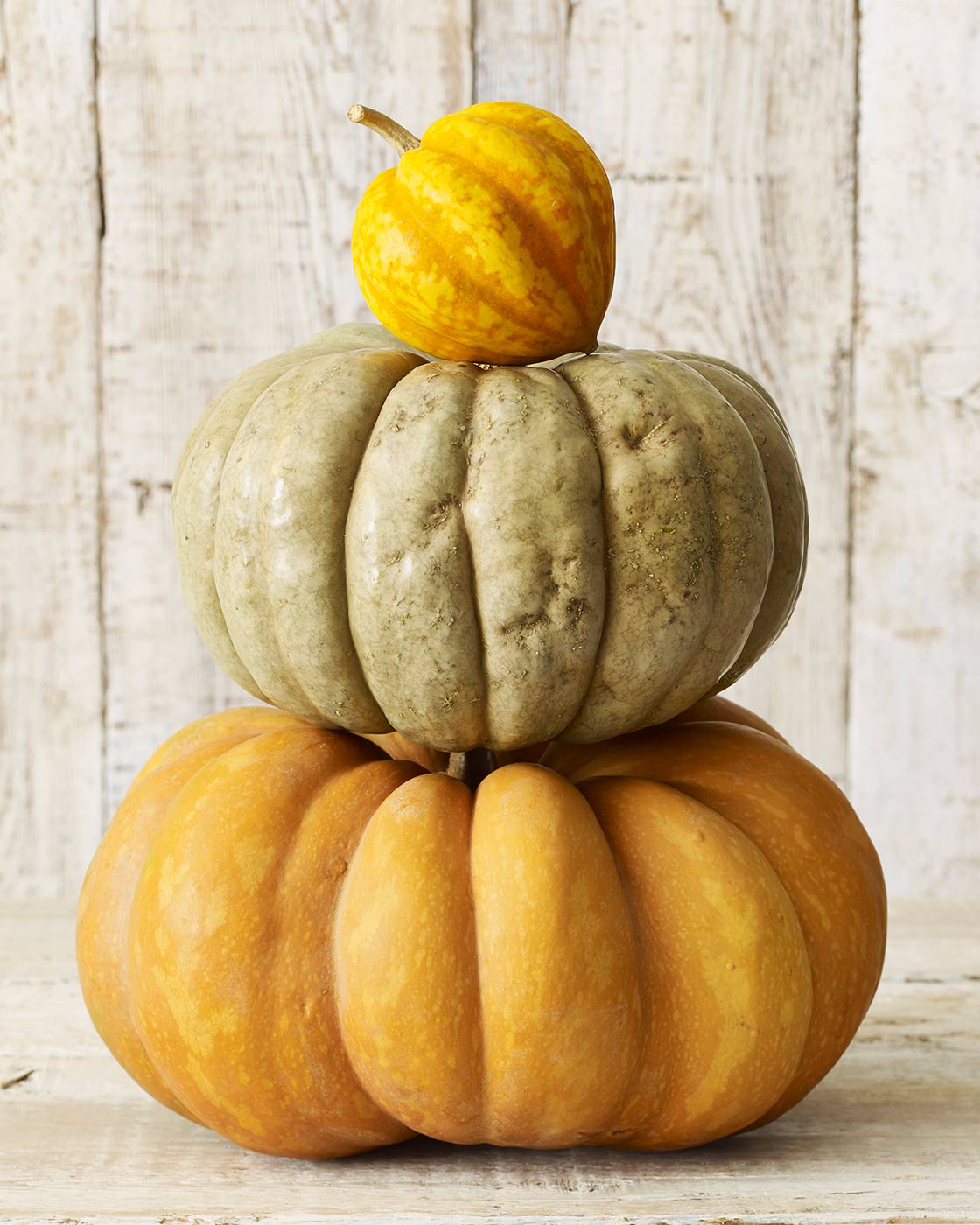 Three stacked pumpkins on a wooden surface with a light wood panel background