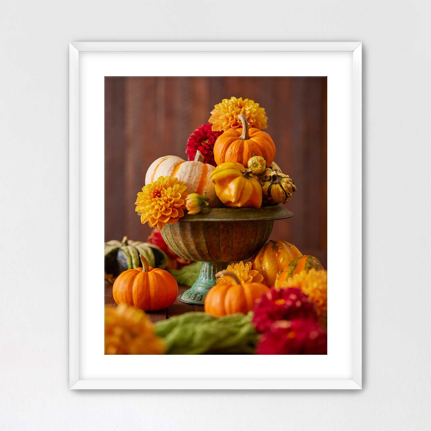 White framed print with a white mat of a still life arrangement with pumpkins and dahlias on a wooden surface.