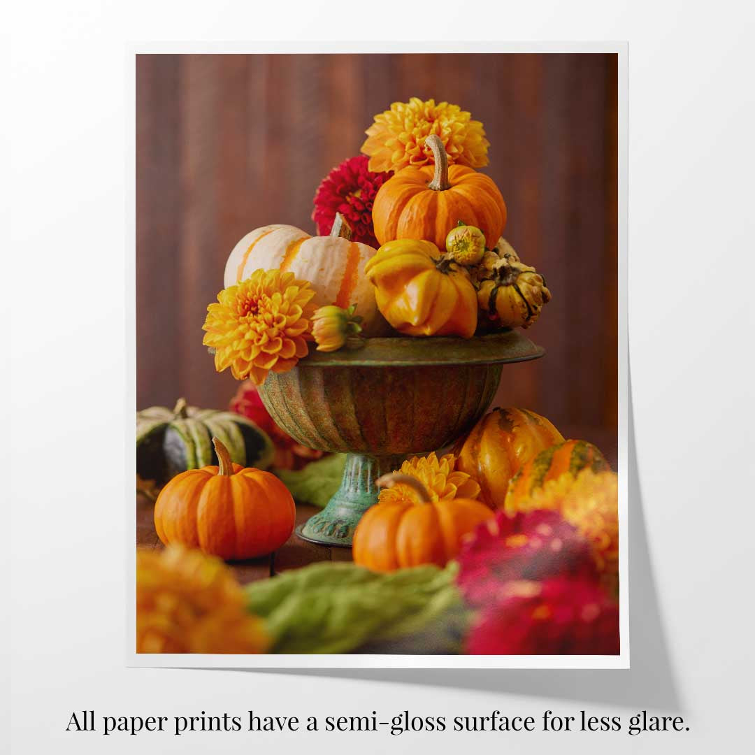 Paper print with a white border of a decorative arrangement of pumpkins and dahlias on a pedestal with a brown background.