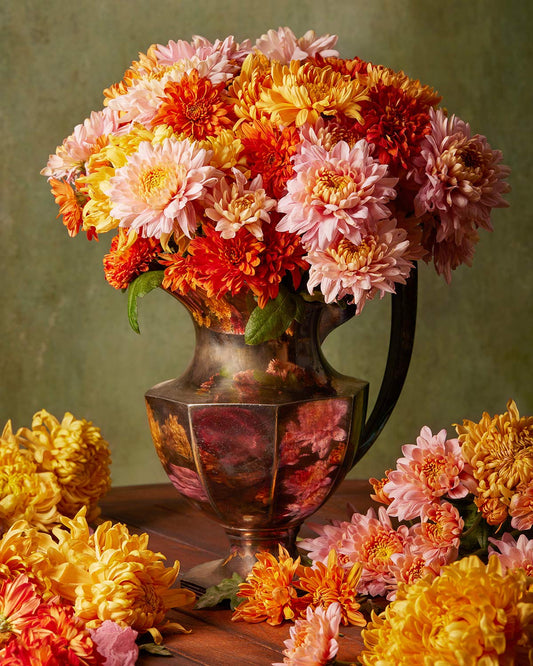 The Harvested Harmony features a antique silver pitcher filled with vibrant pink, orange, and yellow chrysanthemums, exuding the charm of autumn. Additional blooms are scattered on a wooden surface against a muted green background.