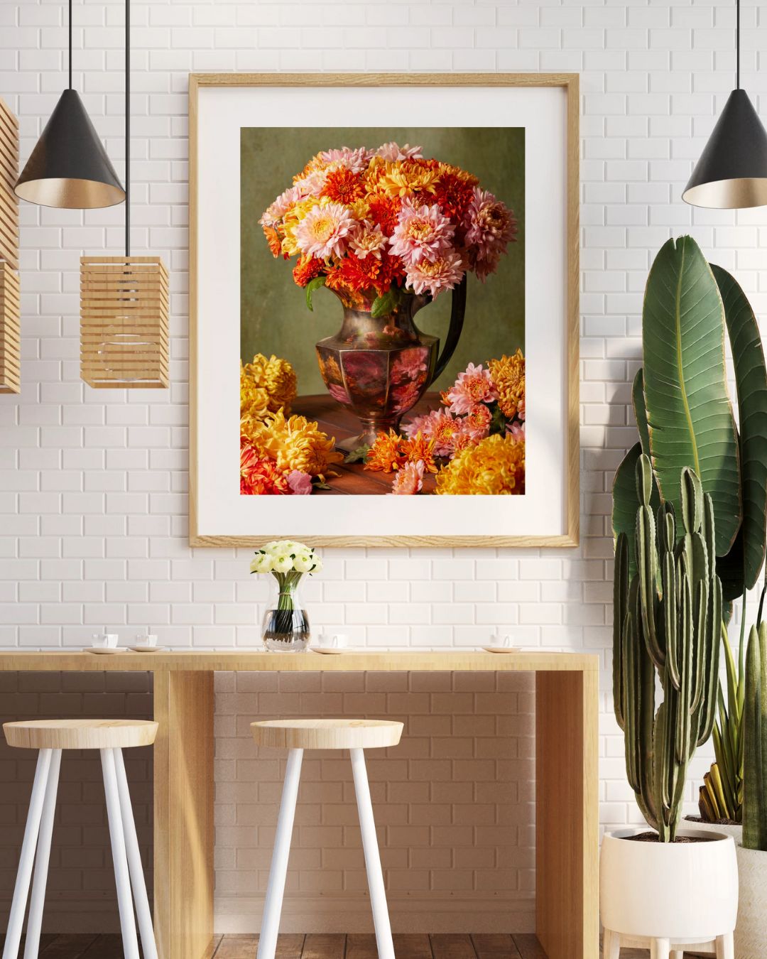 A modern dining area showcases two white stools and a wooden table. A Harvested Harmony fine art print of vibrant autumn flowers hangs above. The backdrop features a white brick wall, pendant lights, a potted plant, and minimalist decor.